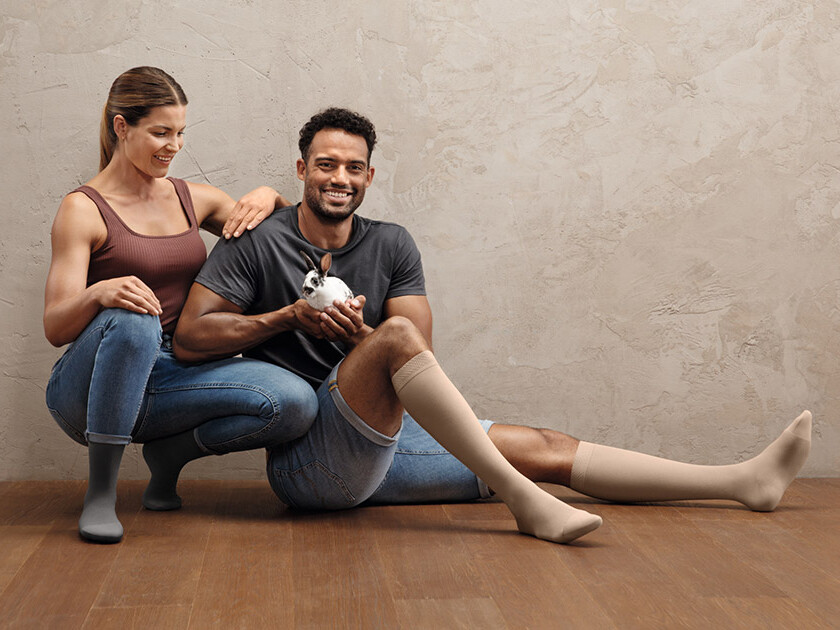 Una pareja de aspecto deportivo está sentada en el suelo. Ambos llevan medias de compresión Juzo Intenso. El hombre sostiene un conejito en la mano. Una pareja de aspecto deportivo está sentada en el suelo. Ambos llevan medias de compresión Juzo Intenso. El hombre sostiene un conejito en la mano.