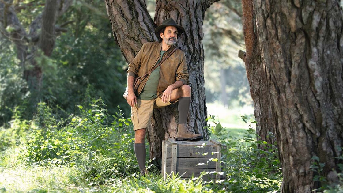 Ein Mann mit Entdecker-Outfit steht in der Natur und schaut in die Ferne. Er trägt kniehohe Kompressionsstrümpfe.