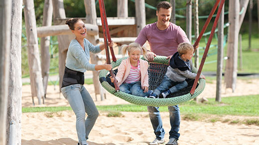 Eine Familie auf dem Spielplatz. Die Kinder schaukeln. Die Mutter trägt eine Rückenbandage Eine Familie auf dem Spielplatz. Die Kinder schaukeln. Die Mutter trägt eine Rückenbandage