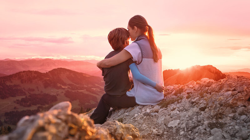 Eine Mutter sitzt mit ihrem Kind auf einem Berggipfel. Das Kind trägt einen Kompressionsärmel. Eine Mutter sitzt mit ihrem Kind auf einem Berggipfel. Das Kind trägt einen Kompressionsärmel.